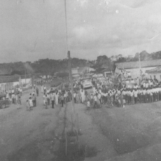 Imagem estilo paisagem. É possível ver uma grande faixa de terra batida e ao centro várias pessoas e pequenas casas.