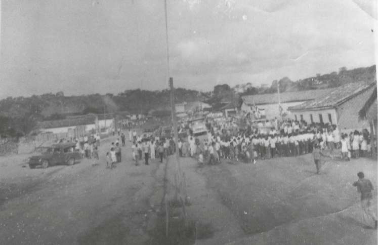 Imagem estilo paisagem. É possível ver uma grande faixa de terra batida e ao centro várias pessoas e pequenas casas.
